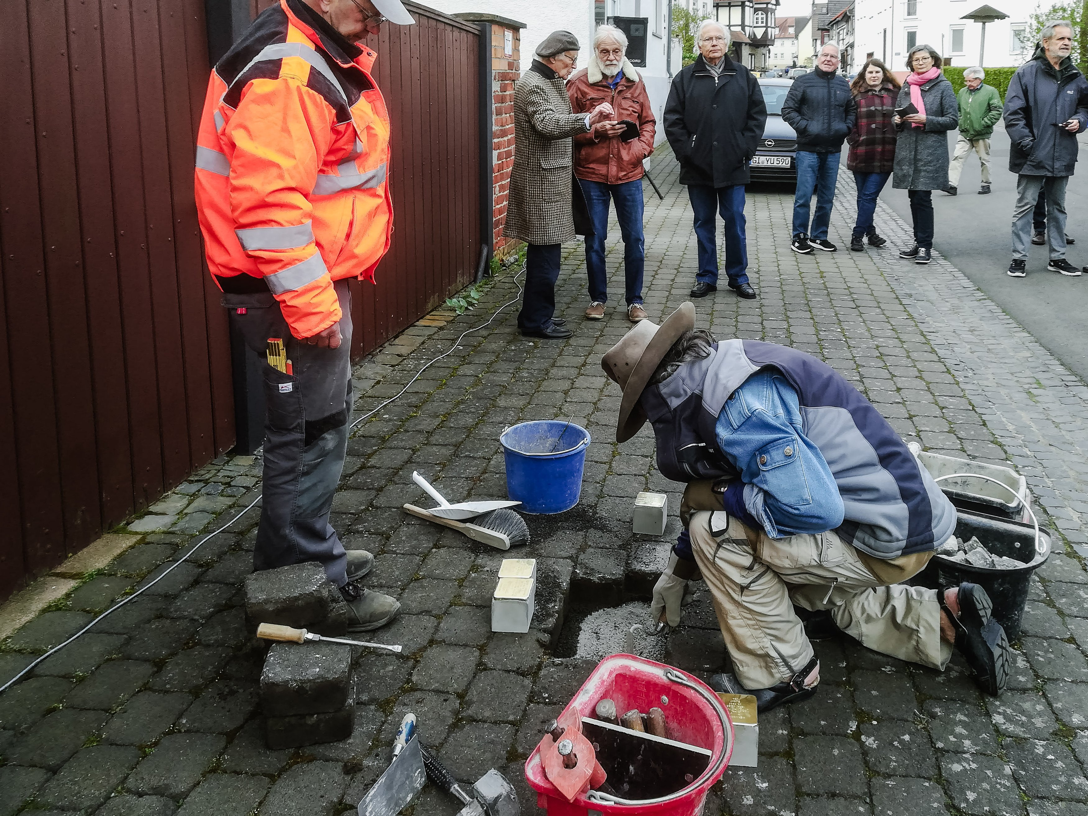 Ein Mann mit Hut kniet am Gehweg und setzt Steine, dahinter Zuschauer
