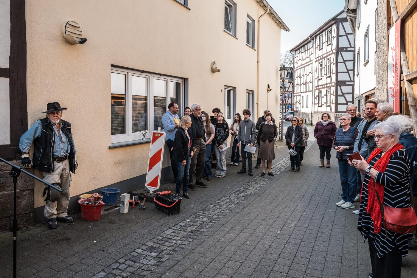 Menschen stehen im Halbkreis in einer Gasse mit Fachwerkhäusern; links kniet Gunter Demnig an der Hauswand.