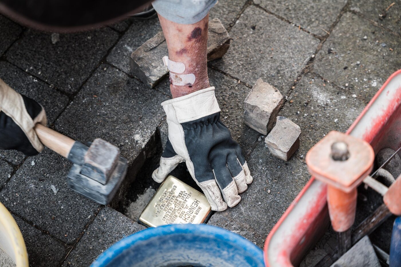 Eine Hand im Arbeitshandschuh setzt einen Stolperstein in die Lücke im Pflaster; daneben Hammer und Eimer.