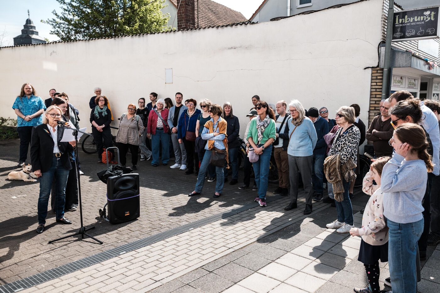 Viele Menschen stehen im Halbkreis vor einer weißen Mauer; eine Frau spricht ins Mikrofon.