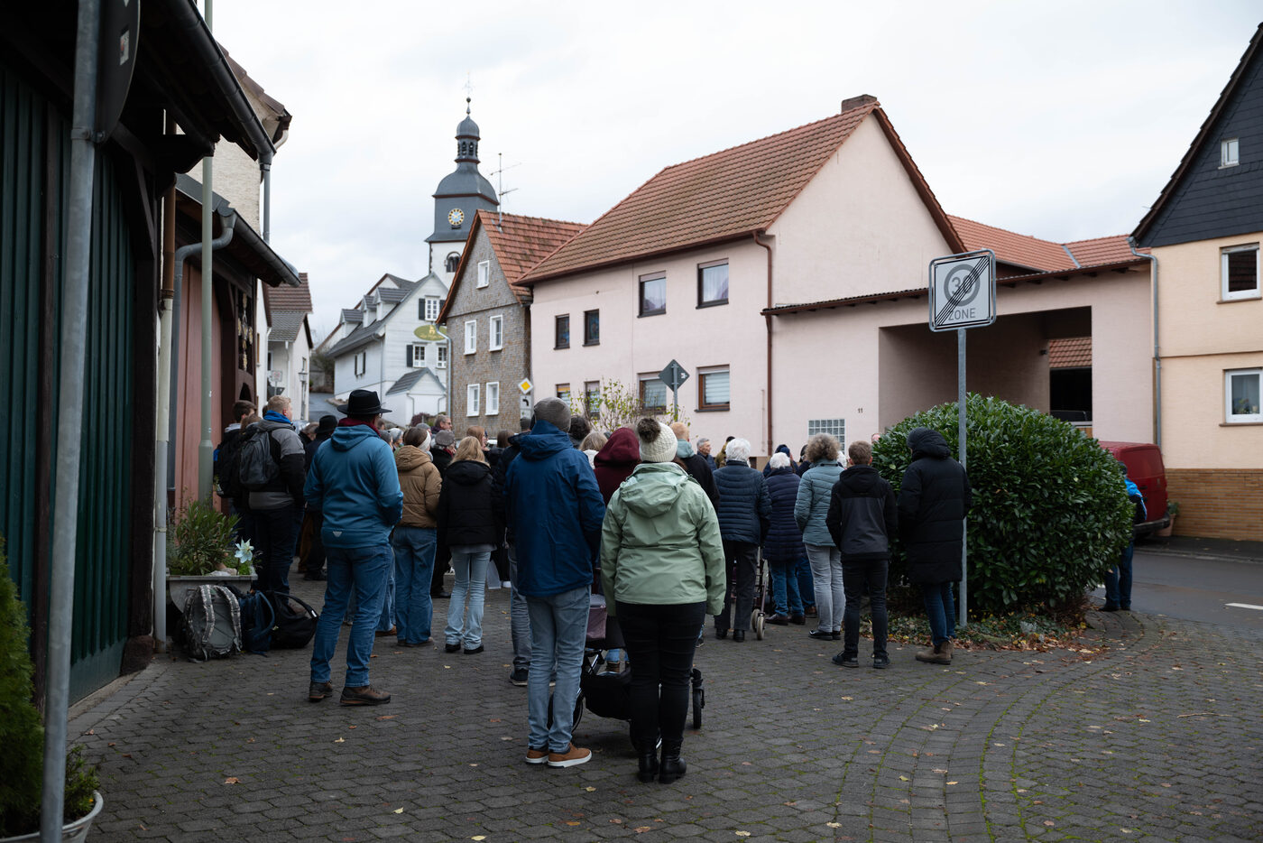Menschen stehen auf einem Platz im Dorf, im Hintergrund Wohnhäuser und ein Kirchturm.