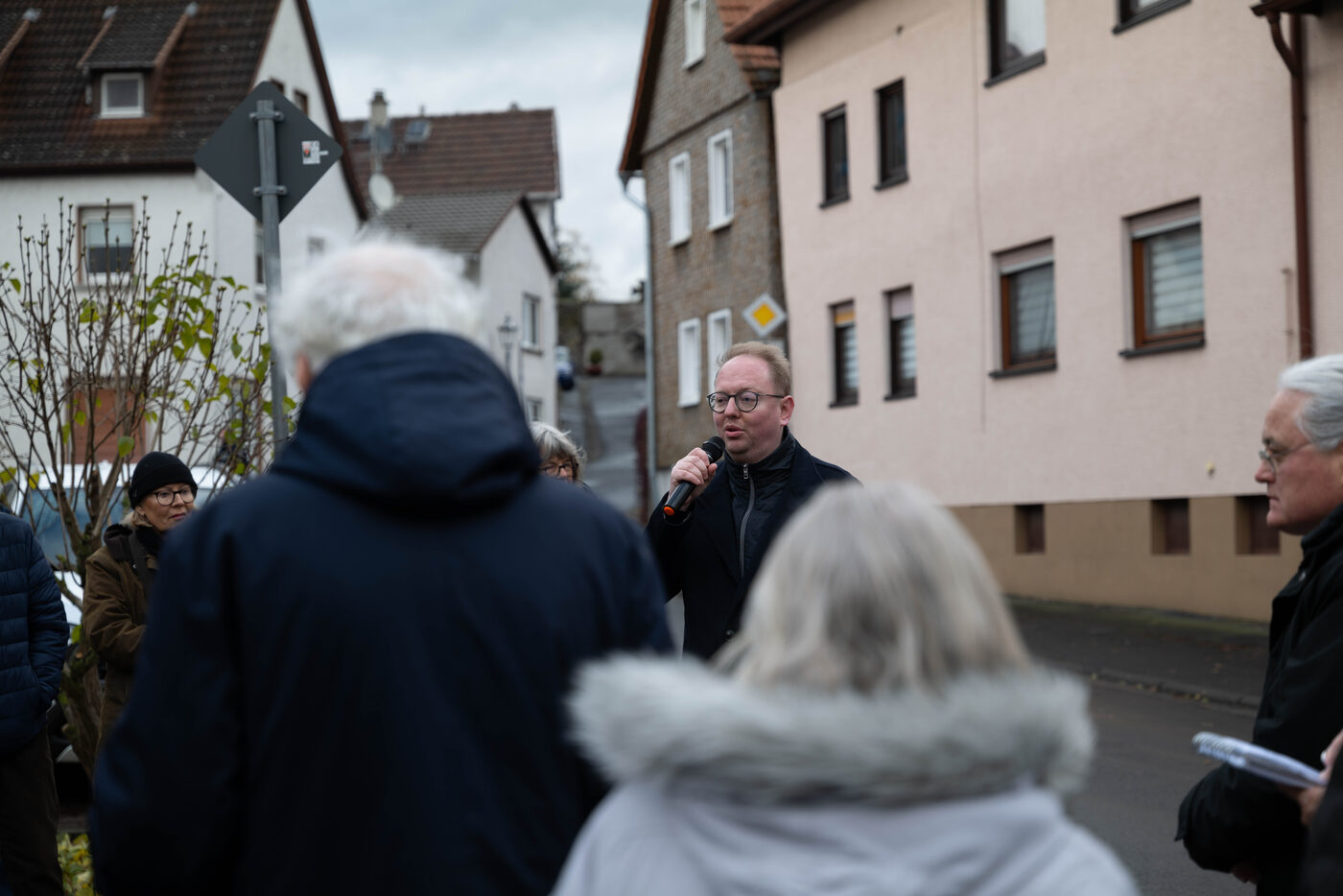 Ein Mann spricht ins Mikrofon, im Vordergrund Zuhörende von hinten.