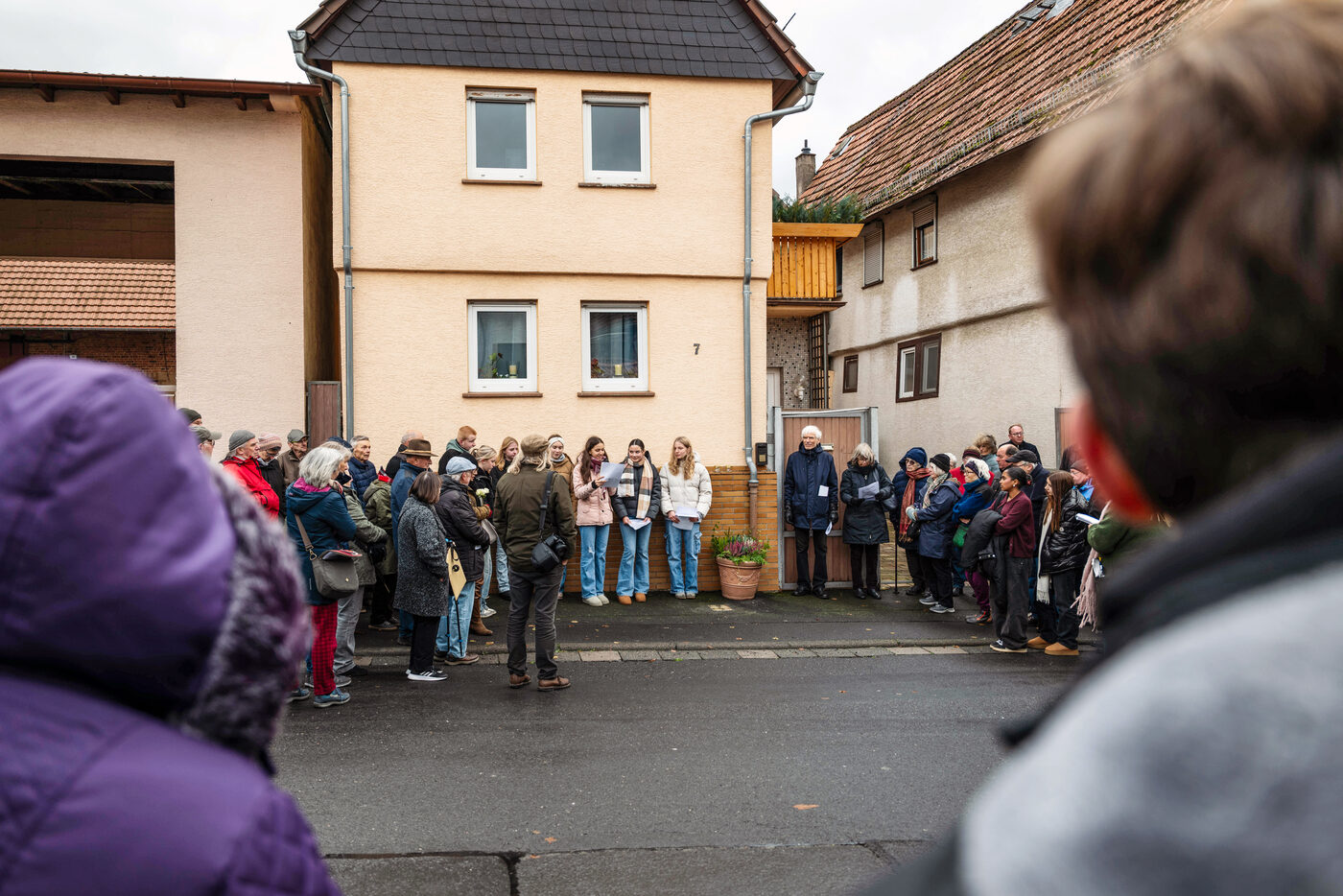 Zuhörende von hinten, gegenüber eine Reihe junger Leute vor einem Wohnhaus.