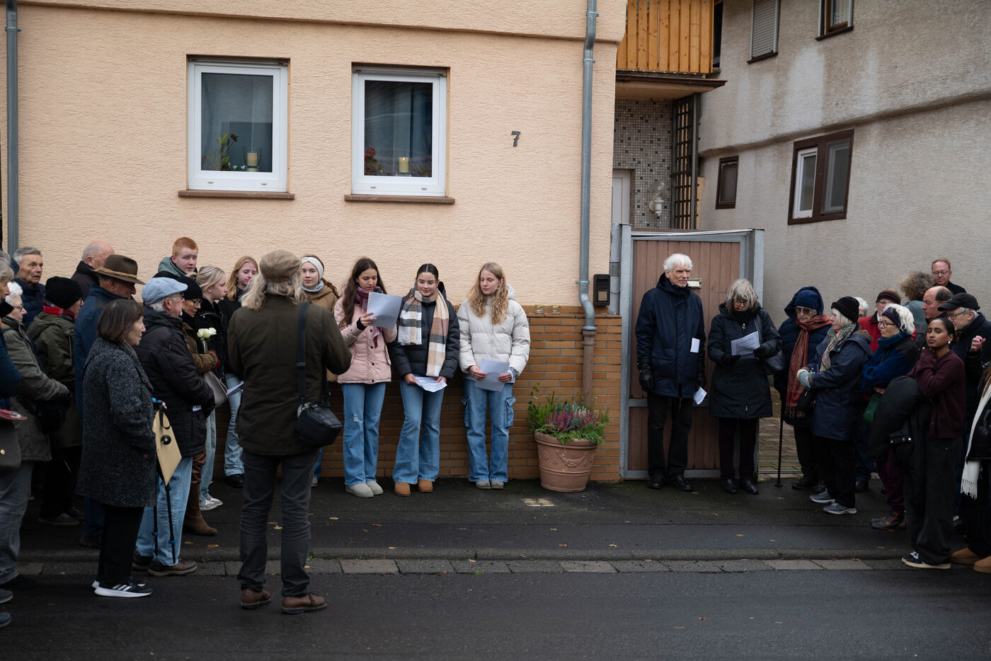 Jugendliche stehen mit Textblättern vor einem Haus mit der Nummer 7, ringsum Zuhörende.