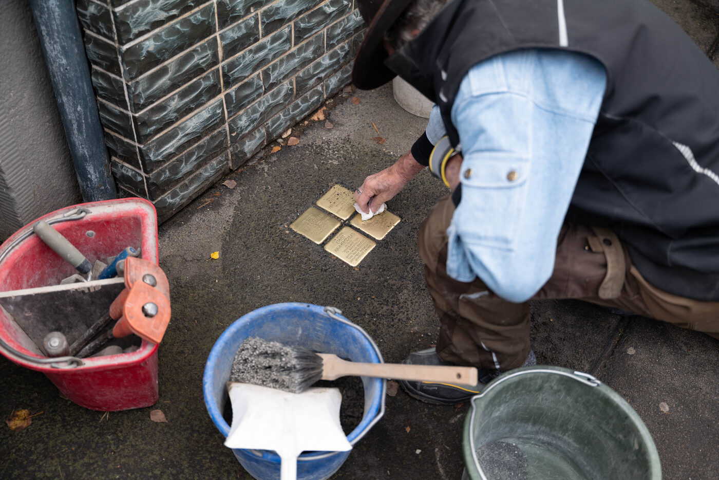 Gunter Demnig kniet an einer Hauswand und richtet vier Stolpersteine aus; daneben Eimer und Handfeger.