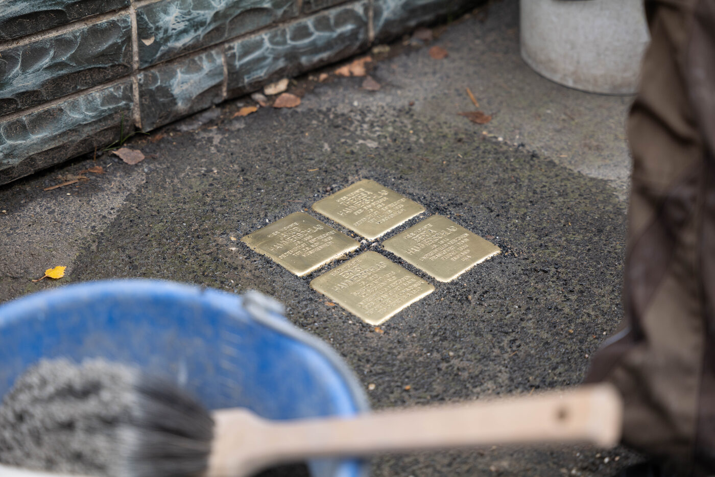 Vier frisch verlegte Stolpersteine im Asphalt, vorn ein Eimer mit Handfeger.