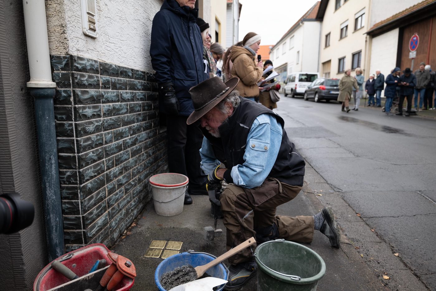 Gunter Demnig mit Hut kniet an einer Hauswand über vier frisch gesetzten Stolpersteinen; im Hintergrund Zuschauer auf der Straße.