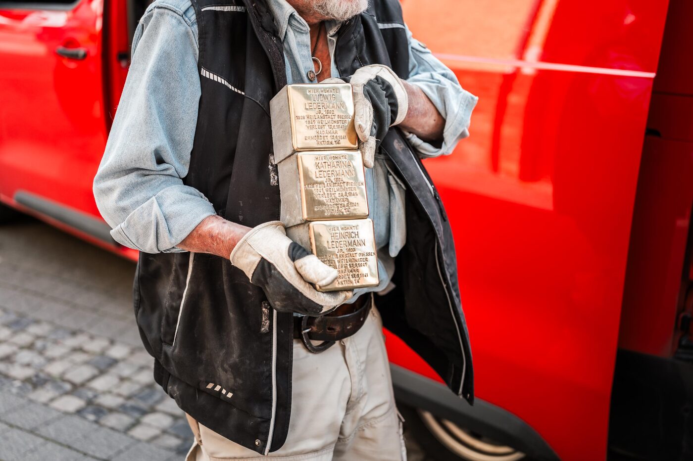 Gunter Demnig trägt drei Stolpersteine im Arm, hinter ihm sein roter Transporter.