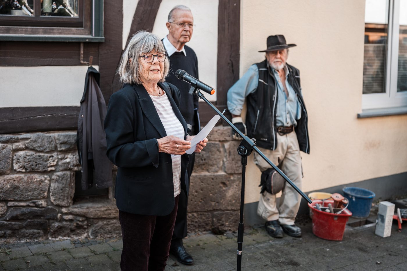 Eine Frau spricht ins Mikrofon, neben ihr ein Mann und Gunter Demnig vor einer Hauswand.