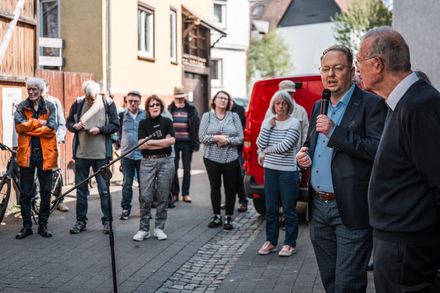 Ein Mann spricht ins Mikrofon, vor ihm stehen die Gäste im Halbkreis in der Gasse.