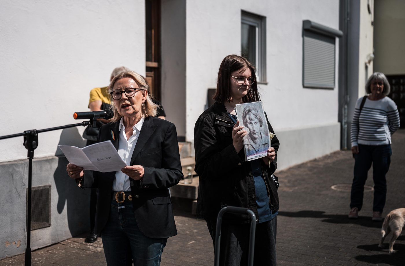 Zwei Frauen am Mikrofon: Die eine liest vom Blatt, die andere hält ein Porträtfoto.