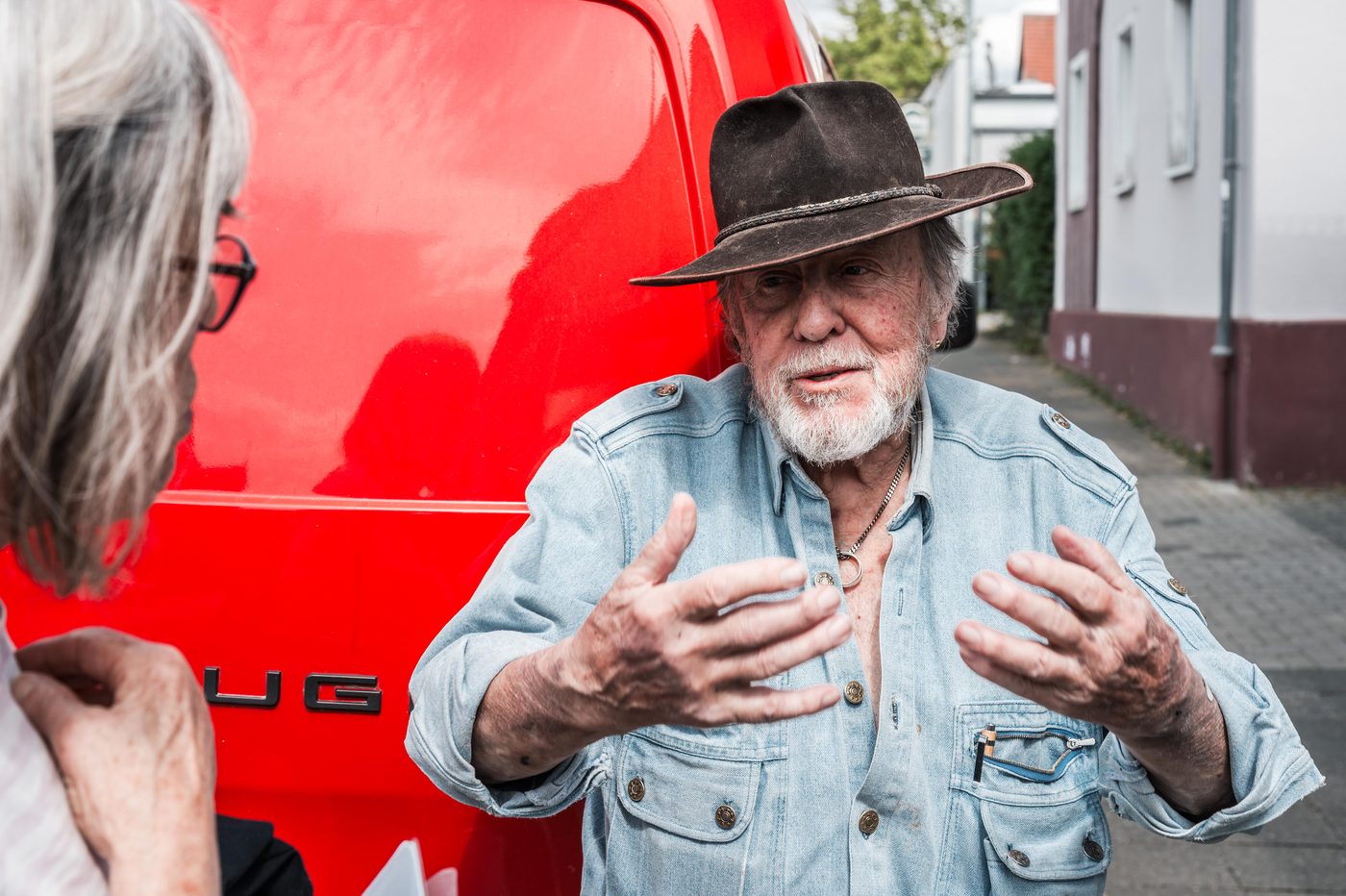 Gunter Demnig mit Hut spricht mit erhobenen Händen vor seinem roten Transporter.